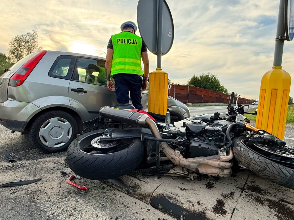 Policjant w kamizelce odblaskowej bada miejsce wypadku, gdzie przewrócony motocykl leży obok uszkodzonego auta.