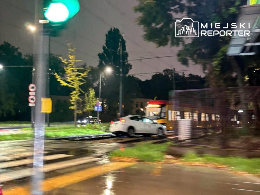 Biały samochód osobowy stoi na torowisku tramwajowym w deszczowej noc, otoczony przez latarnie i zieleń.