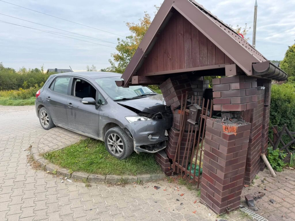 Samochód osobowy w kolorze szarym wjeżdża w murowaną wiatę śmietnikową, uszkadzając jej strukturę.