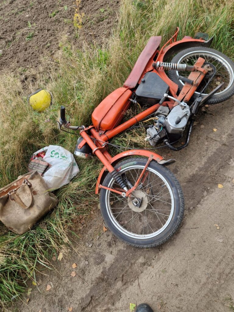 Czerwony motorower leży na poboczu drogi, obok znajduje się torba i żółta kask. W tle widoczna uprawa.