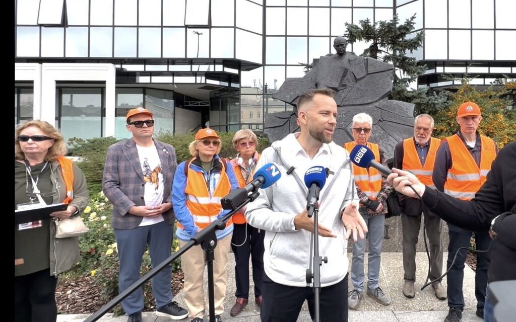 Mężczyzna w szarej bluzie przemawia do mikrofonu, a za nim grupa osób w pomarańczowych kamizelkach.