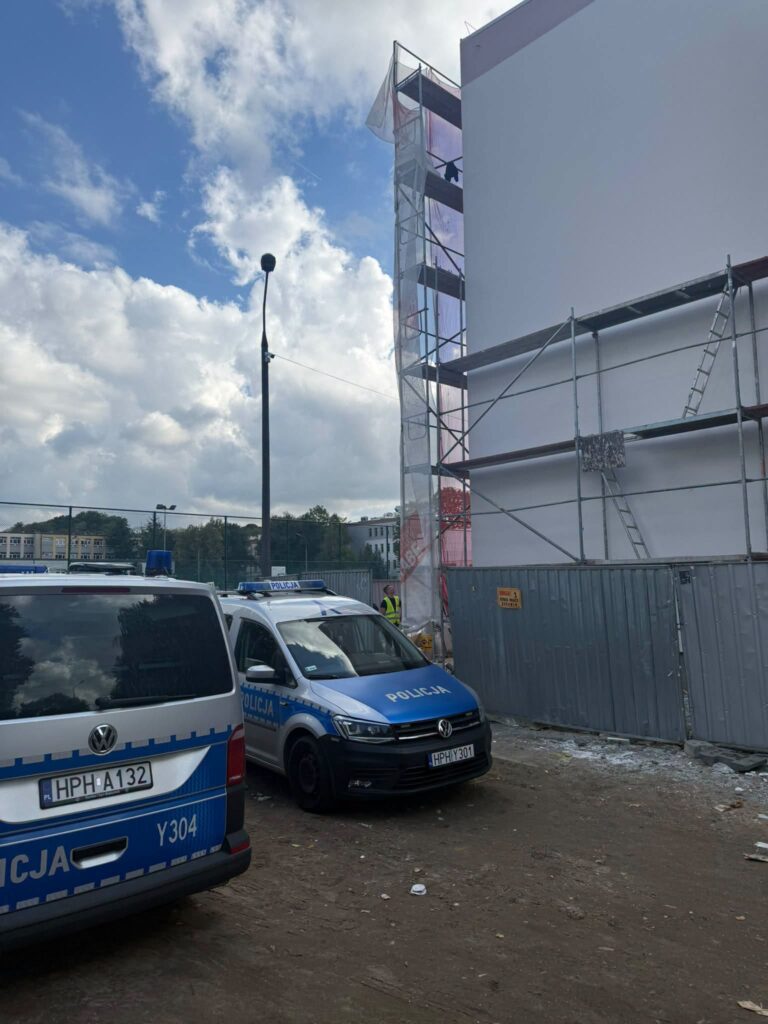 Policyjne pojazdy zaparkowane obok budowy, na której pracownik znajduje się na rusztowaniu. W tle chmury na niebie.