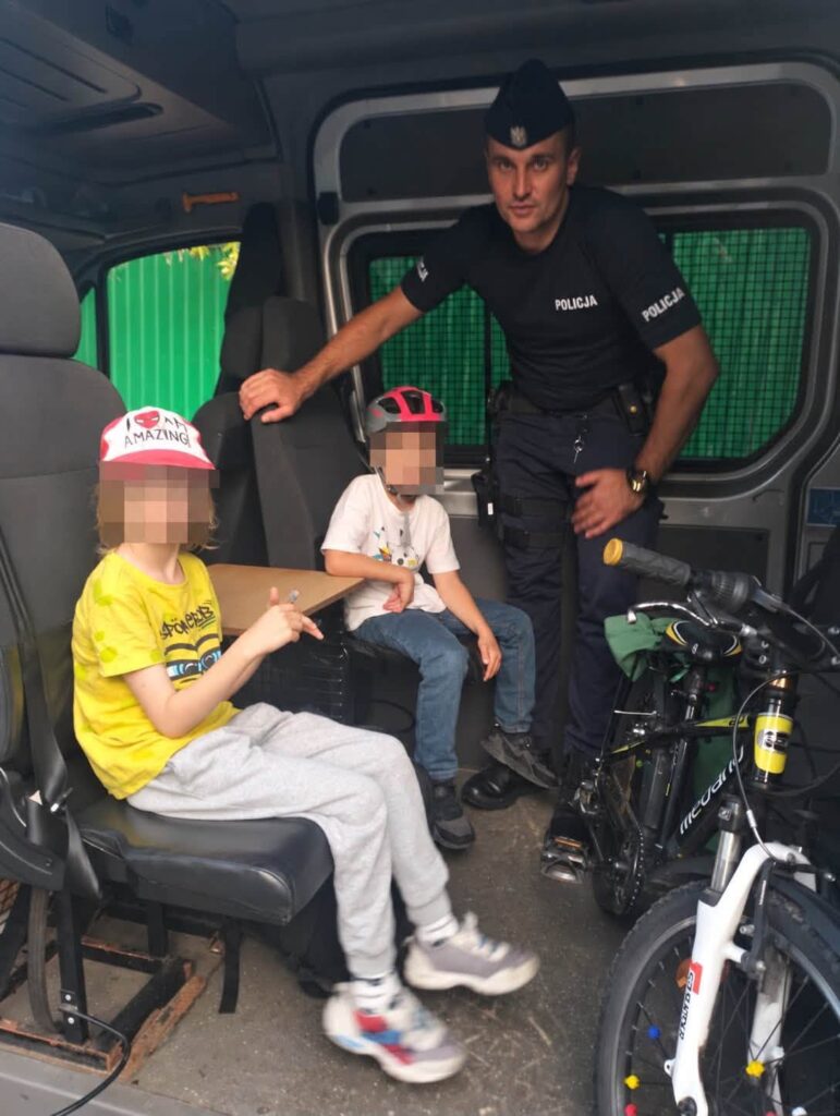 Policjant w mundurze siedzi w samochodzie policyjnym obok dwóch chłopców, którzy mają kaski na głowach.