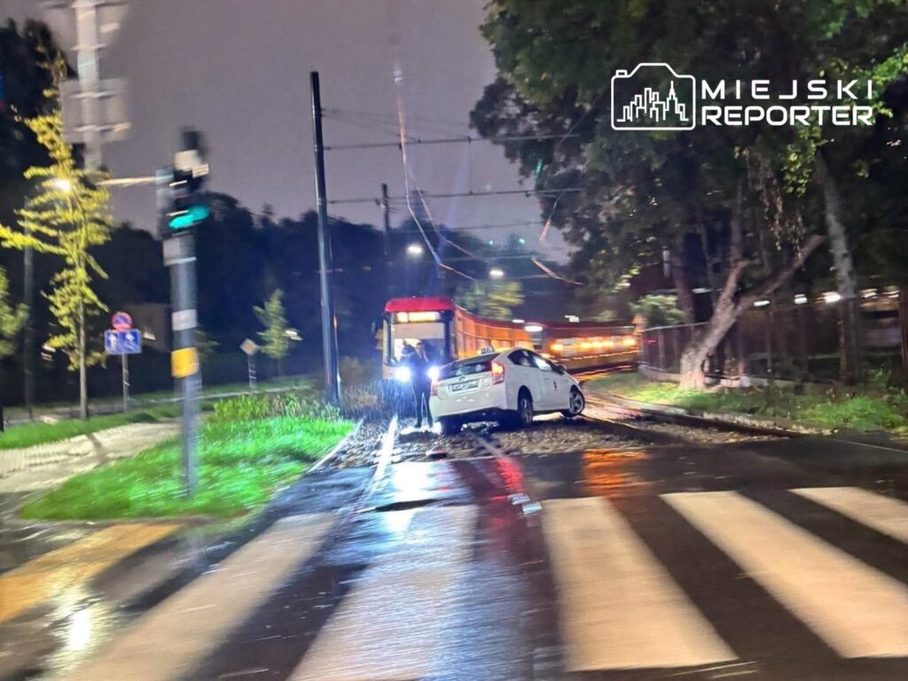Biały samochód osobowy stoi na torach tramwajowych w deszczowym otoczeniu, obok przejścia dla pieszych.