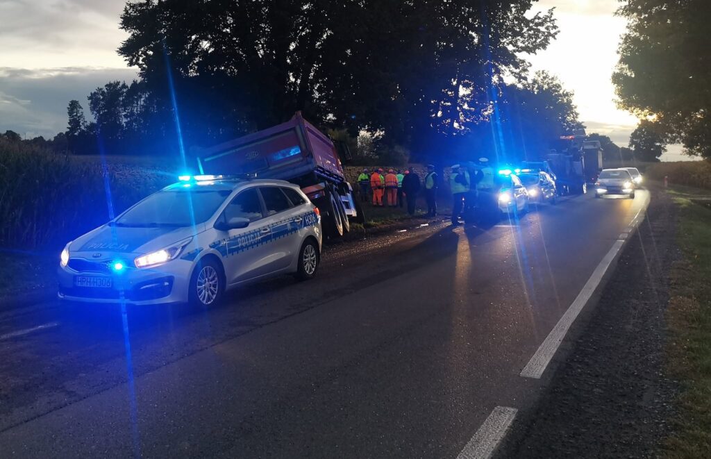 Na drodze stoi wywrócona ciężarówka, a obok grupka osób w odblaskowych kamizelkach rozmawia z policjantami.