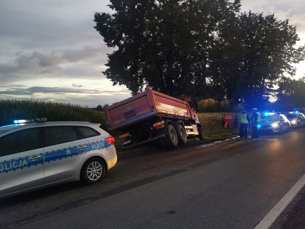 Czerwony samochód ciężarowy leży na boku przy drodze, obok grupy policjantów rozmawiających z mężczyzną.