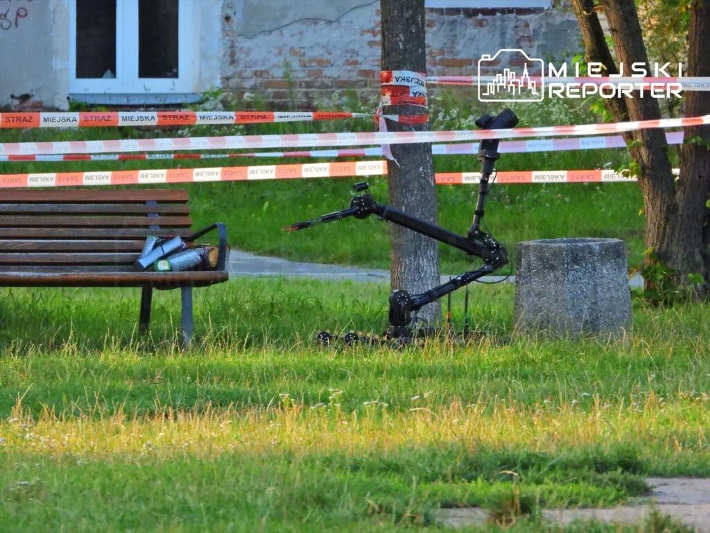 Robot z ramieniem mechanicznym umieszczony obok betonowego słupa, w otoczeniu trawnika i ławeczki, otoczony taśmą ostrzegawczą.