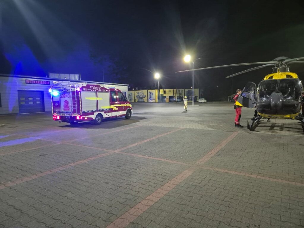 Śmigłowiec ratunkowy stoi na nocnym parkingu obok wozu strażackiego i ratowników.