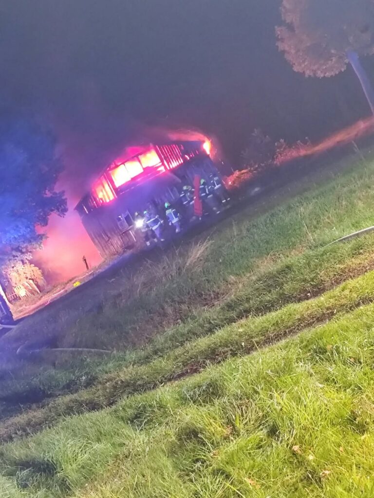 Strażacy gaszą płonący drewniany budynek nocą; z dachu wydobywają się płomienie i gęsty dym.