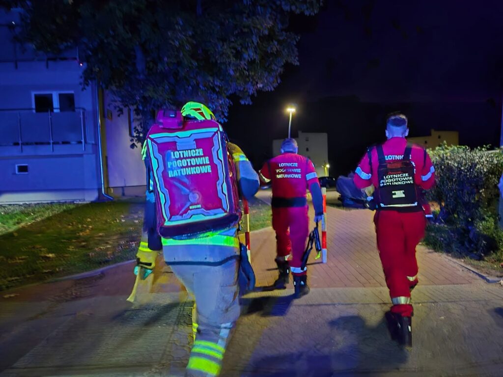 Ratownicy Lotniczego Pogotowia Ratunkowego idą nocą chodnikiem między blokiem a żywopłotem.