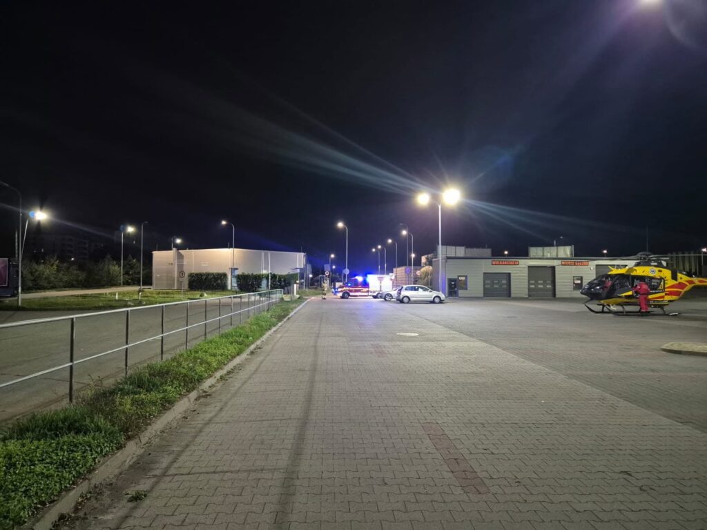 Żółto-czerwony śmigłowiec ratunkowy stoi nocą na placu przy budynku, obok zaparkowanych samochodów ratunkowych.