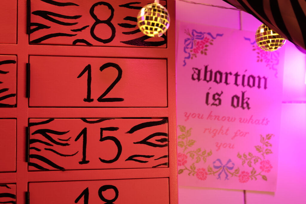 Różowa ściana z ponumerowanymi szufladami i plakatem z napisem „abortion is ok” w świetle dyskotekowych kul.
