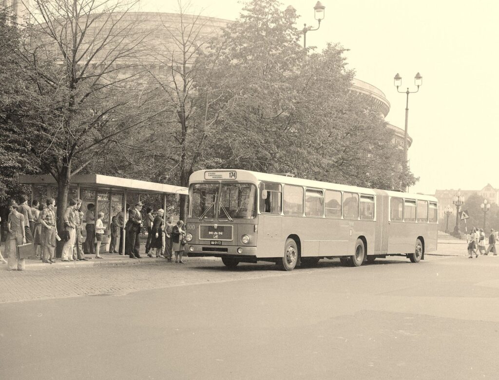 Autobus przegubowy zatrzymuje się na przystanku, a pasażerowie czekają w kolejce w otoczeniu drzew i budynków.