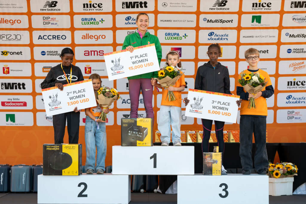 Zwyciężczynie maratonu stoją na podium, trzymając nagrody: pierwsza z kwiatami i czekiem na 15 000 USD, druga z czekiem na 7 500 USD, trzecia z czekiem na 5 000 USD.
