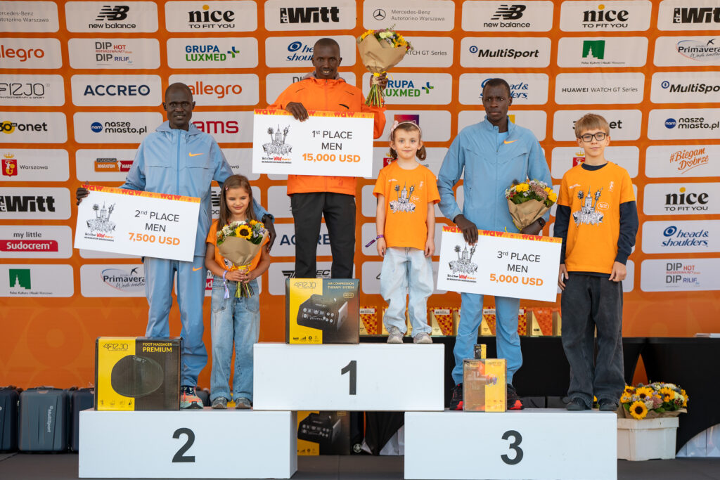 Zawodnicy stoją na podium po maratonie, trzymając nagrody i kwiaty. Pierwsze miejsce zajmuje mężczyzna w pomarańczowej koszulce, drugi z niebieską, a trzeci w czarnej. Obok nich stoją dzieci w pomarańczowych koszulkach.