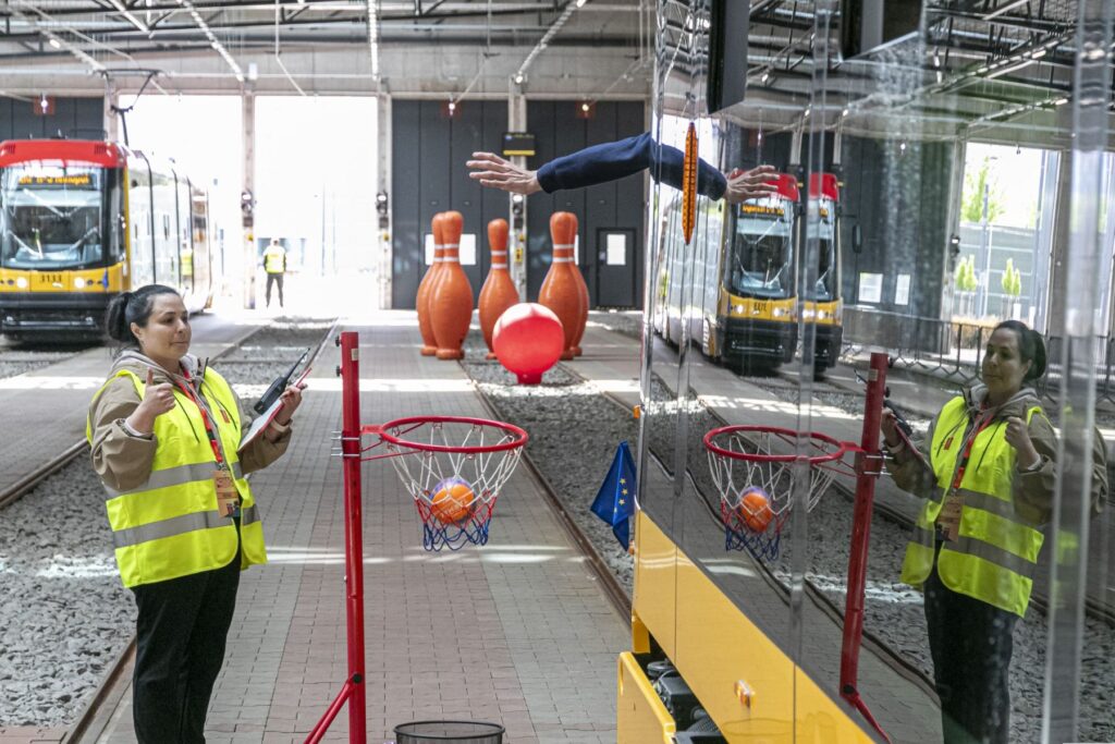 Kobieta w kamizelce odblaskowej trzyma notatnik i wskazuje na kosz do koszykówki w zajezdni tramwajowej.