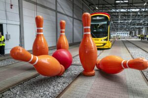 Wielkie, pomarańczowe kręgle oraz czerwona kula leżą na torach tramwajowych w nowoczesnym zajezdni, obok nadjeżdżającego tramwaju.