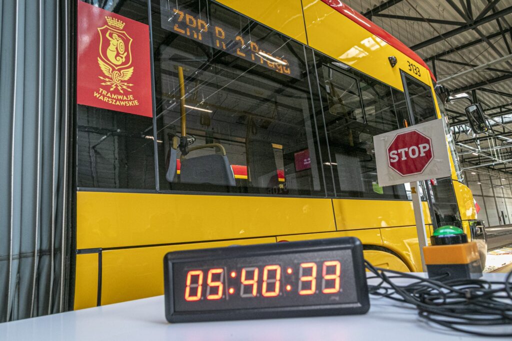 Żółty tramwaj z logo "Tramwaje Warszawskie" stoi w zajezdni, a przed nim widoczny jest zegar odliczający czas.
