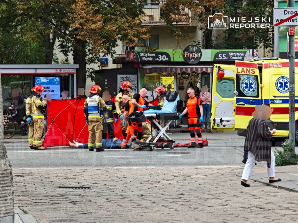 Ratownicy medyczni oraz strażacy udzielają pomocy osobie leżącej na ulicy, obok stoi ambulans.