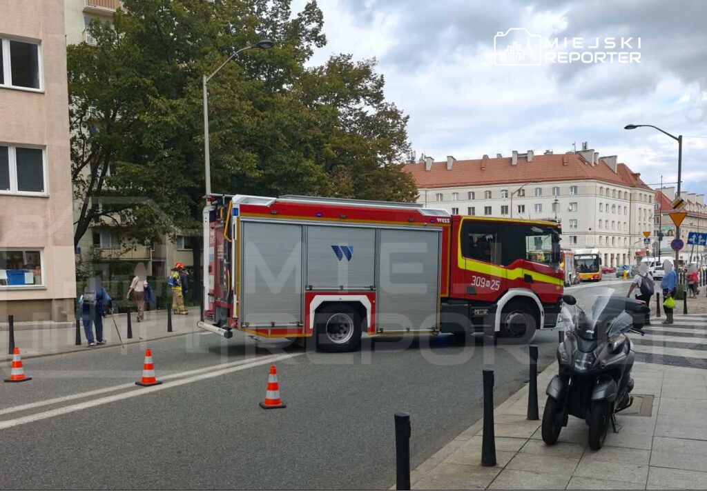 Wozidło strażackie z włączonymi sygnalizacjami zatrzymane przy jezdni, obok przechodniów i budynków mieszkalnych.