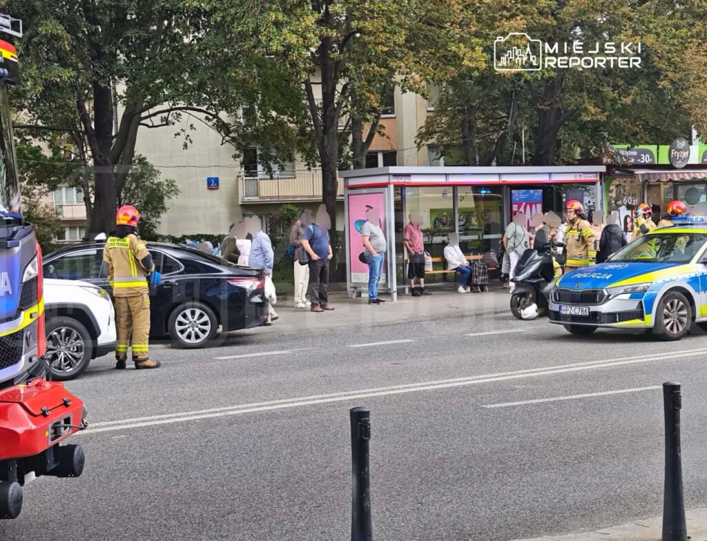 Grupa strażaków i policjantów interweniuje przy czarnym samochodzie osobowym na ulicy, obok przystanku autobusowego.