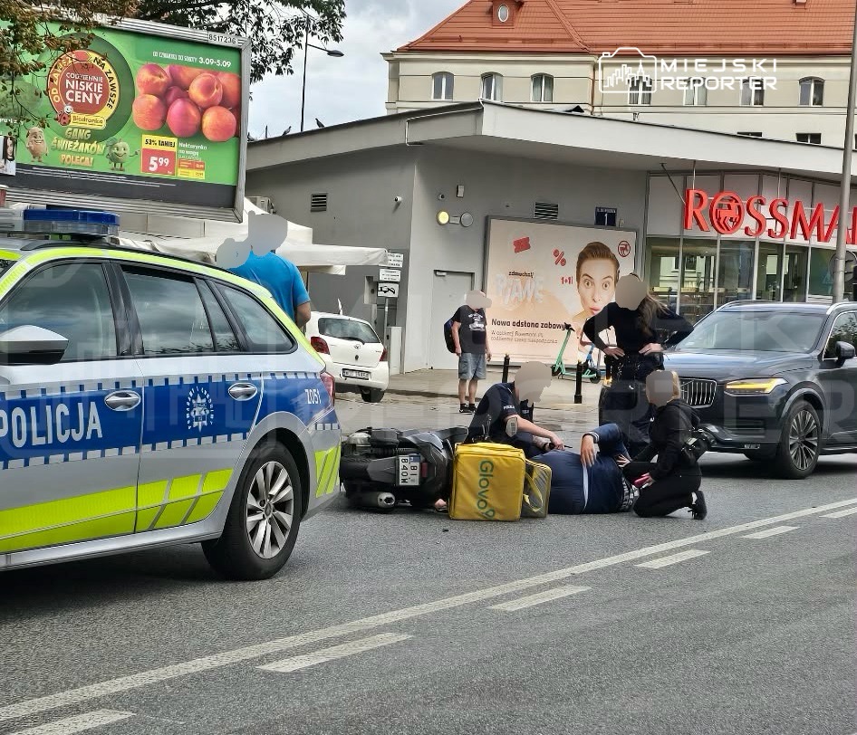 Policjanci zatrzymują mężczyznę leżącego na jezdni obok skuterów dostawczych w miejskiej scenerii.