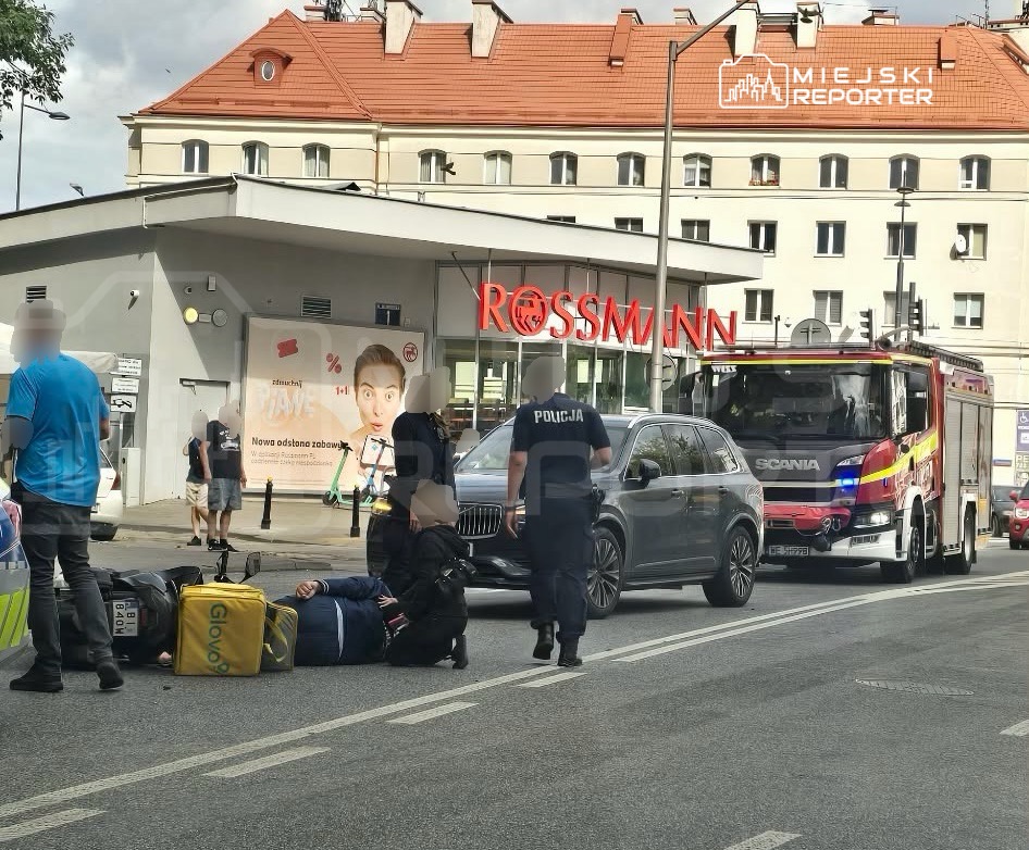 Mężczyzna leżący na jezdni obok żółtej torby dostawczej, otoczony przez policjantów i straż pożarną, w pobliżu sklepu Rossmann.