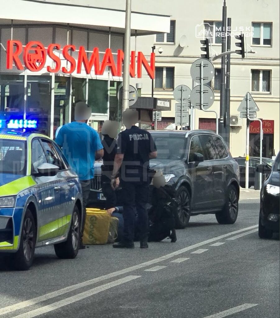 Policjanci rozmawiają z mężczyzną siedzącym na jezdni obok torby dostawczej przed sklepem Rossmann.