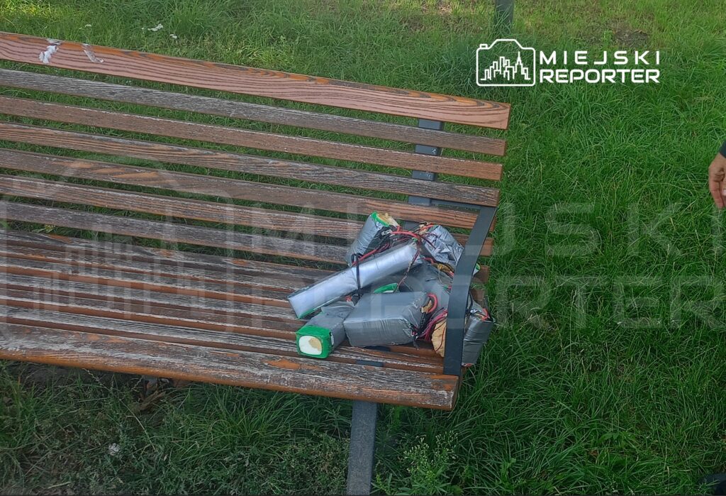 Na ławce w parku leżą przedmioty o metalicznej powierzchni, owinięte taśmą, z widocznymi przewodami.