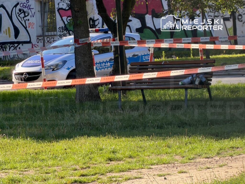 Policyjny radiowóz zaparkowany obok ławeczki, otoczony taśmą zabezpieczającą w zielonym terenie z graffiti w tle.