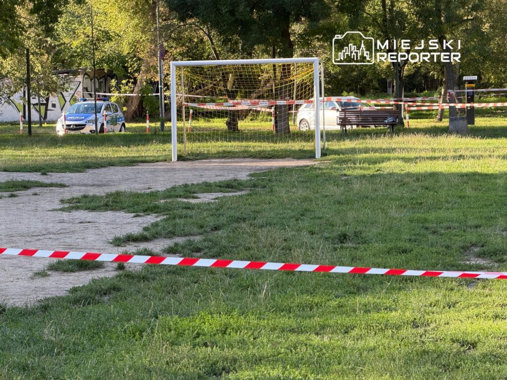 Boisko do piłki nożnej z bramką, otoczone taśmą ostrzegawczą, w tle policyjny samochód i ławka w parku.
