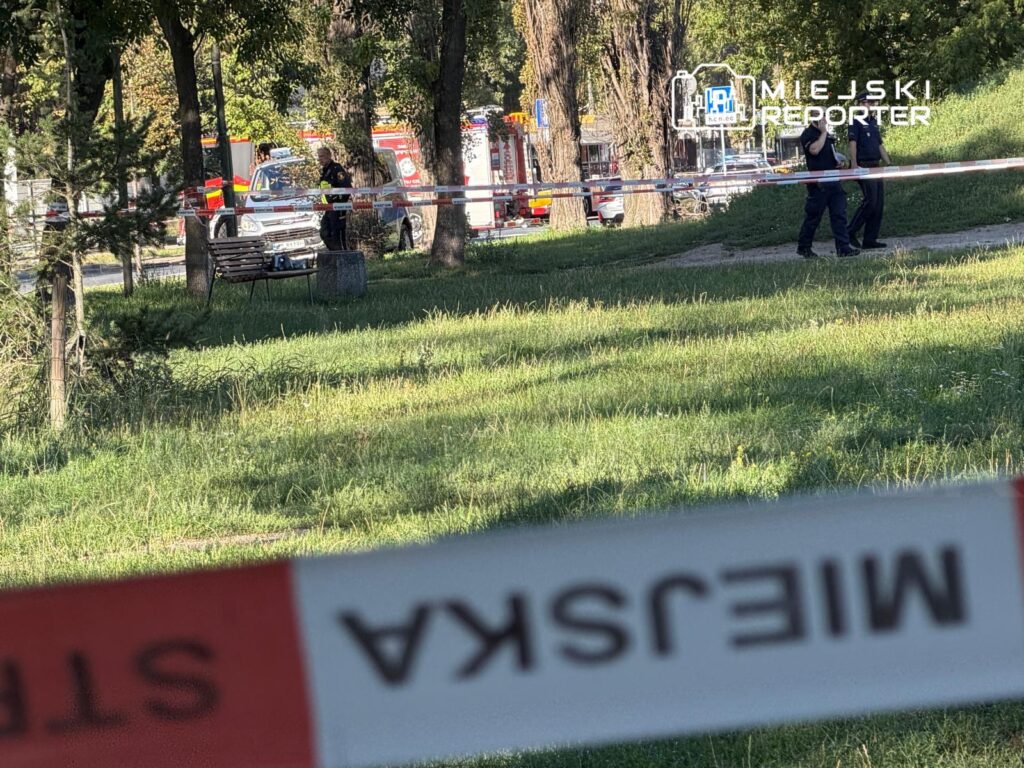 W parku, w tle widać straż pożarną oraz policjantów zabezpieczających teren. Na pierwszym planie znajduje się ławka.