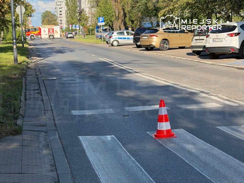 Na jezdni znajduje się pomarańczowy stożek ostrzegawczy, a w tle widoczne są radiowozy i wóz strażacki.