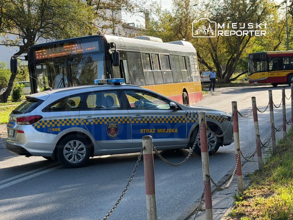 Radiowóz straży miejskiej zatrzymany obok przystanku autobusowego, w tle dwa autobusy komunikacji miejskiej.