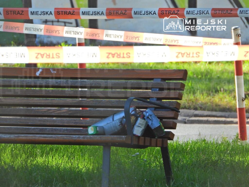 Na ławce w parku leży butelka oraz nieznany przedmiot, obok którego rozciągnięta jest taśma policyjna.