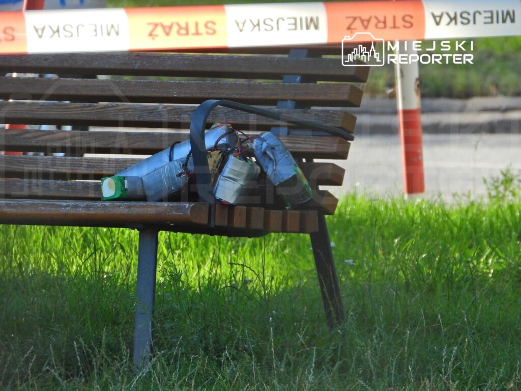 Na ławce w parku leży przedmiot przypominający niebezpieczny ładunek, otoczony taśmą ostrzegawczą.