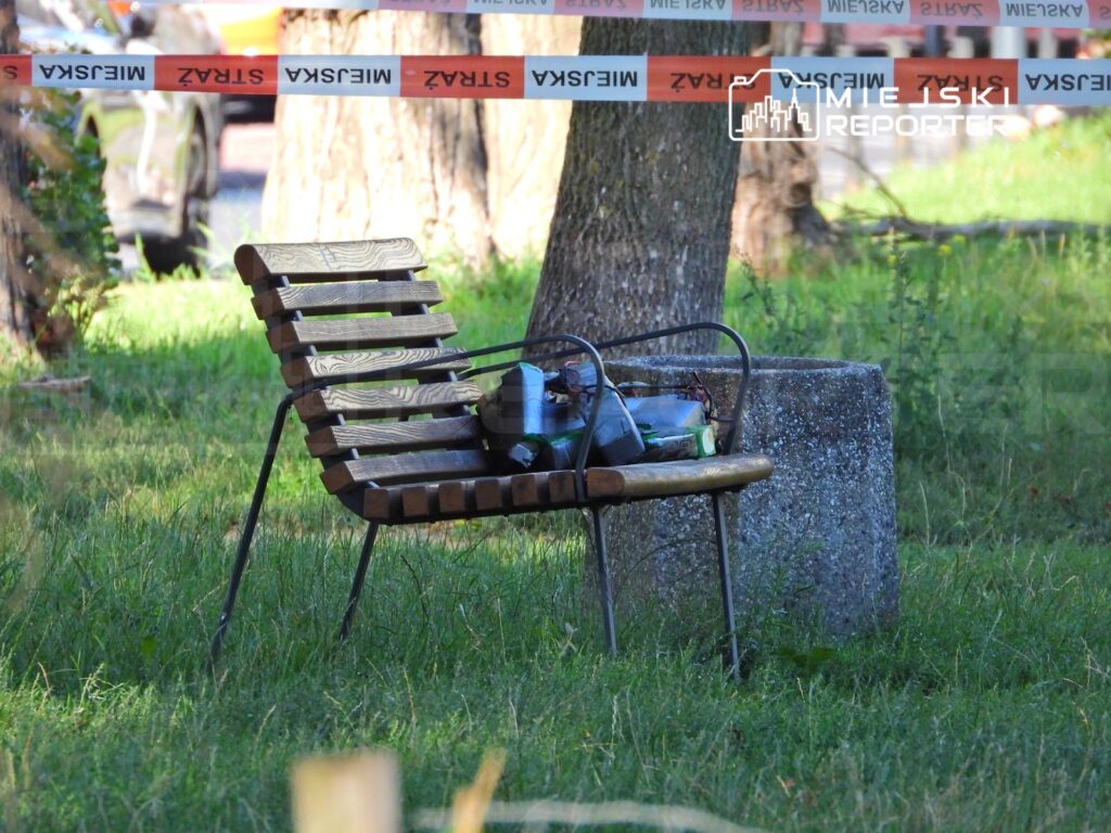Zardzewiały, metalowy stół z drewnianymi siedziskami stoi na trawie, obok niego leży torba z różnymi przedmiotami.