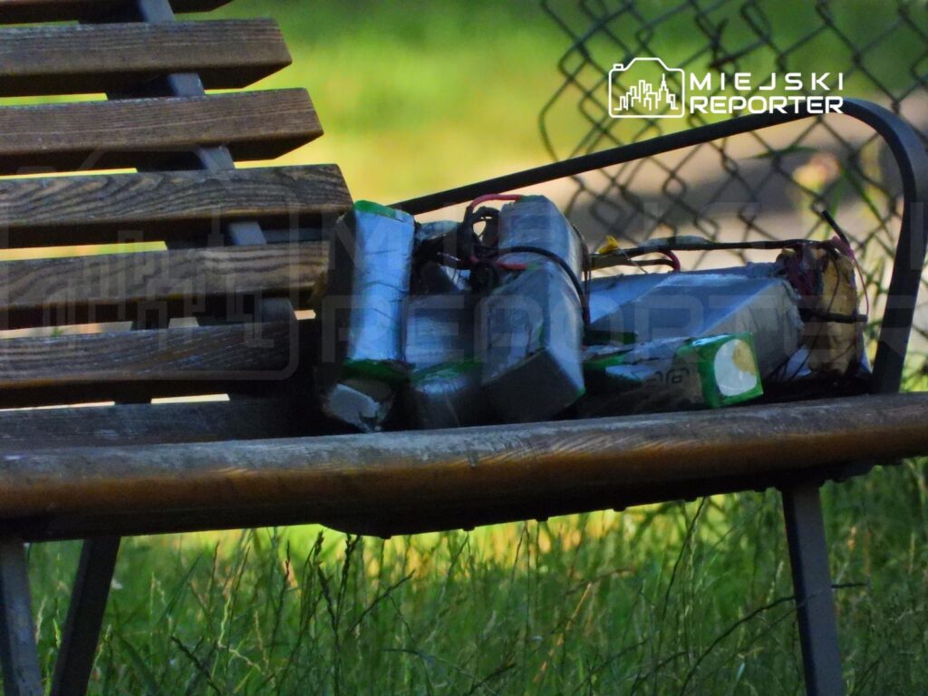 Na drewnianej ławce w parku leżą nieznane przedmioty w opakowaniach, częściowo otwarte, wśród trawy.