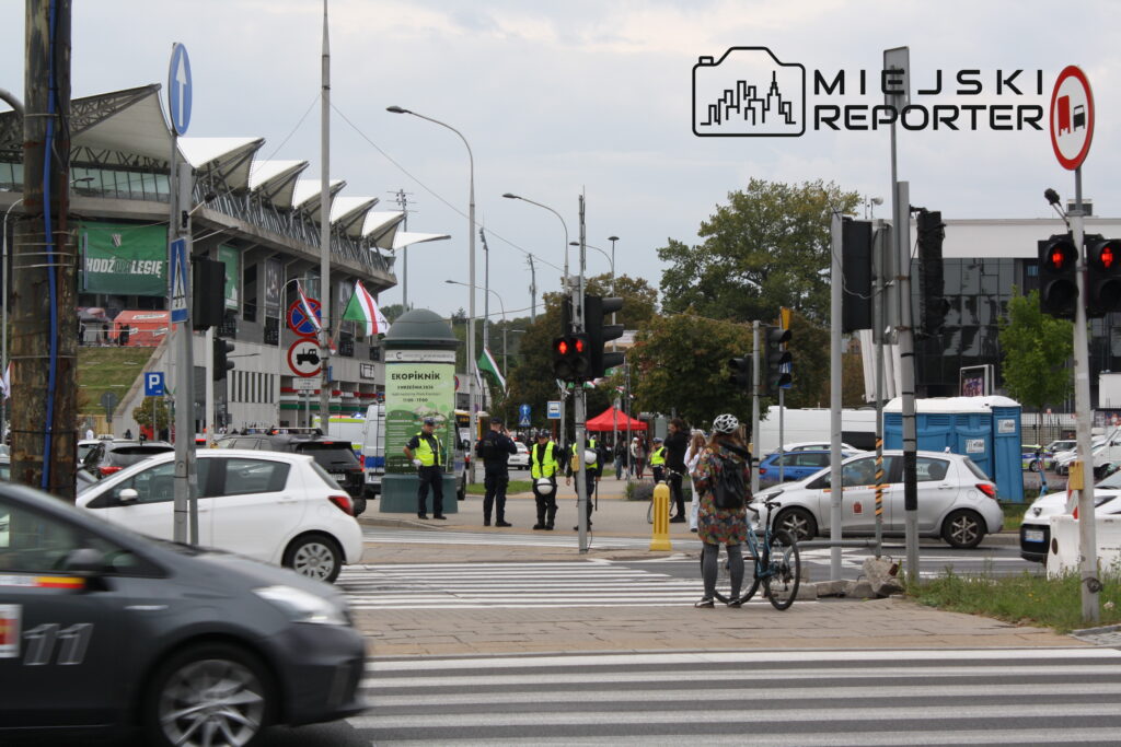 Ruchliwe skrzyżowanie z sygnalizacją świetlną, rowerzystka czeka na zielone światło, w tle stadion i policyjna kontrola.