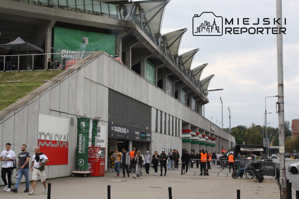 Grupa ludzi przechodzi obok nowoczesnego stadionu, gdzie odbywa się wydarzenie. W tle widać zadaszenie i reklamy.