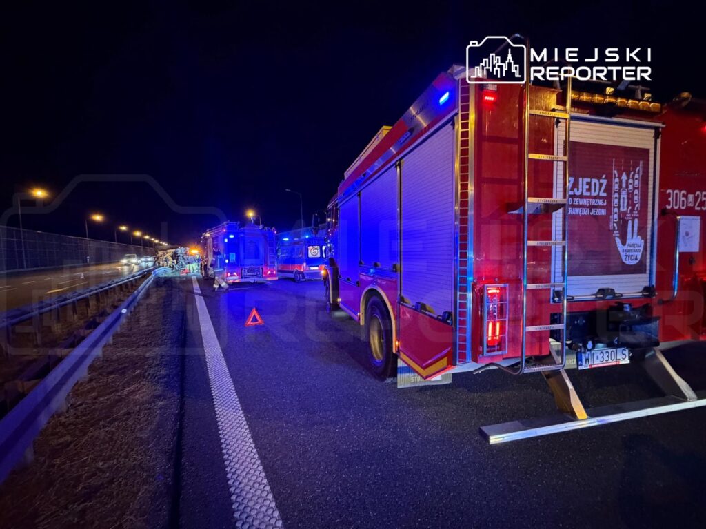 Na drodze nocą stoją wozu strażackie z włączonymi sygnalizatorami, a w tle widoczne są pojazdy.