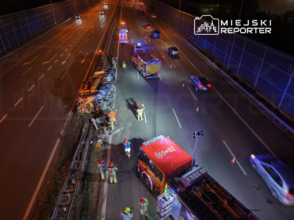 Grupa strażaków pracuje przy przewróconym pojeździe na autostradzie, w tle widać pojazdy ratunkowe i sygnalizację świetlną.