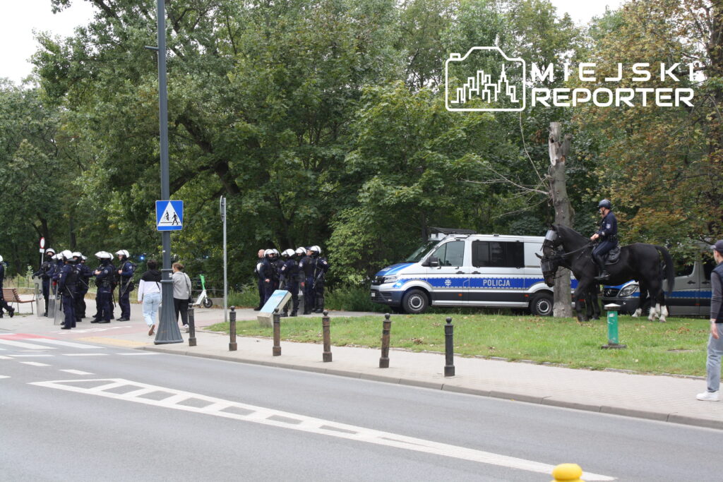Grupa policjantów w mundurach stoi w pobliżu radiowozu, a na koniu patroluje funkcjonariusz.