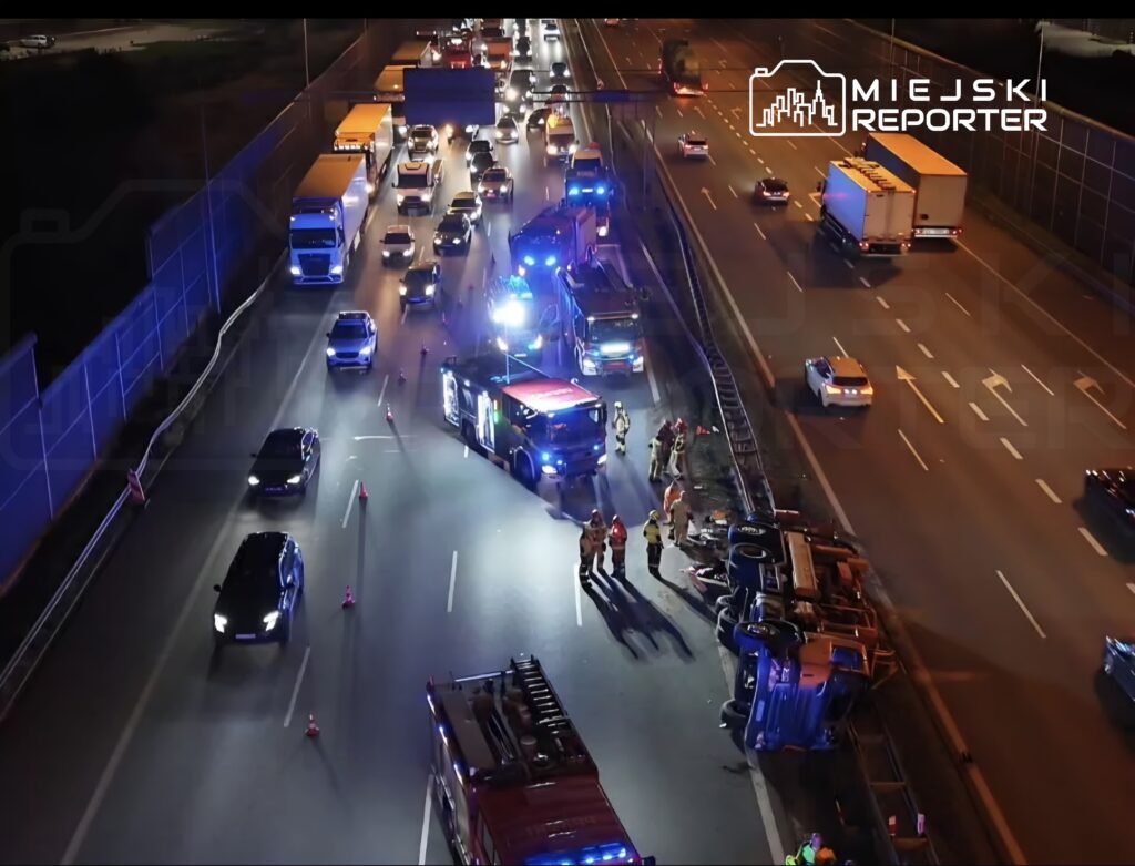 Na ruchliwej drodze nocą, strażacy i służby ratunkowe pracują przy przewróconym pojeździe, a ruch jest kierowany objazdem.
