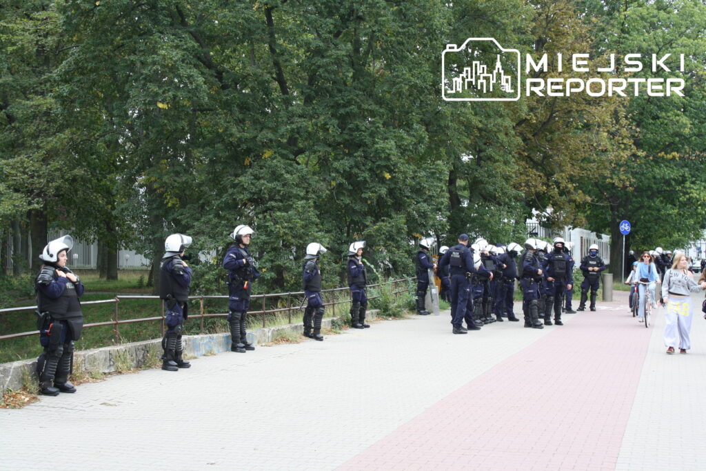 Grupa policjantów w hełmach i mundurach stoi wzdłuż chodnika, podczas gdy przechodnie spacerują obok.