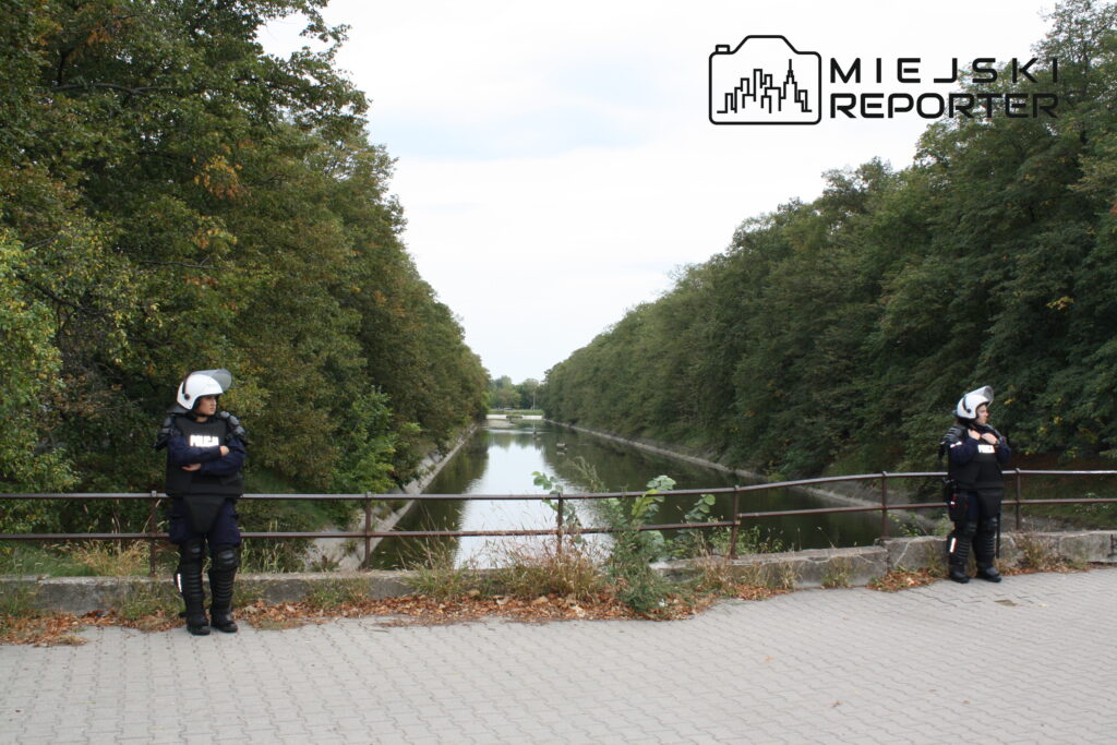 Dwóch policjantów w hełmach stoi na brzegu rzeki, otoczeni przez drzewa i zarośla.