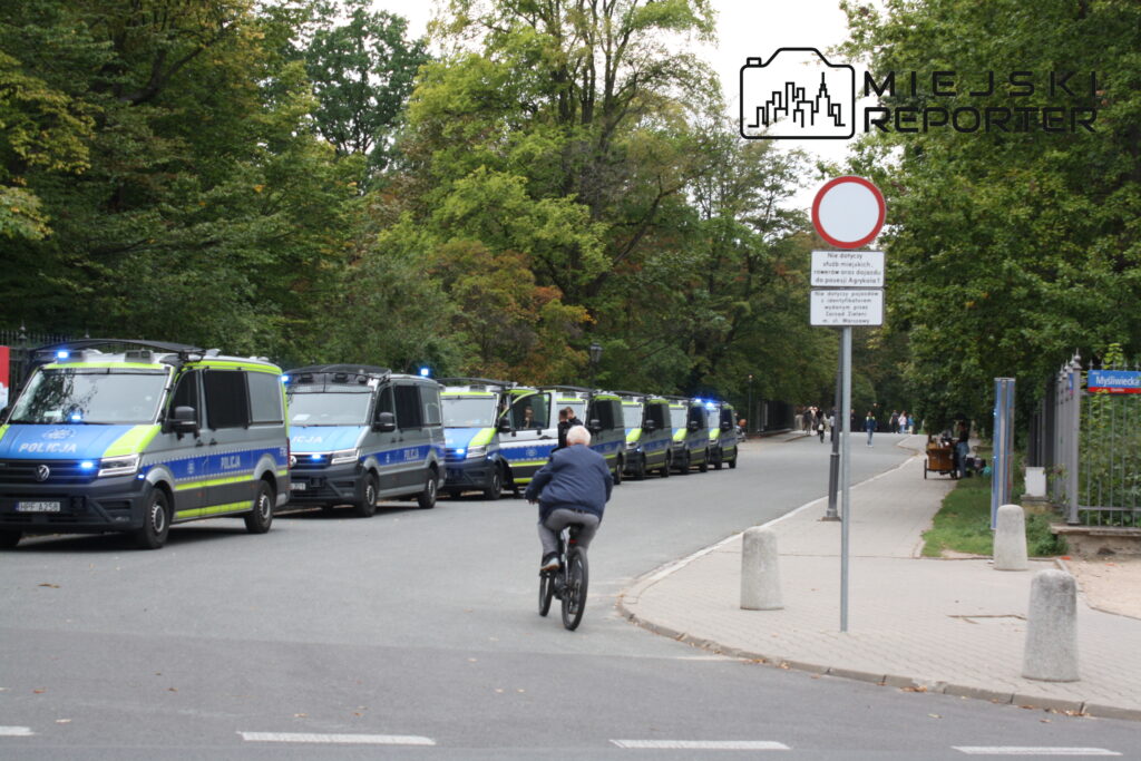 Mężczyzna na rowerze przejeżdża obok linii policyjnych furgonetek zaparkowanych przy drodze w parku.