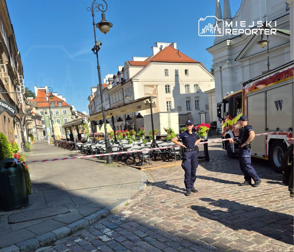 Policyjny patrol zabezpiecza teren z czerwonymi taśmami w pobliżu zamkniętej ulicy, obok wozu strażackiego i restauracji.