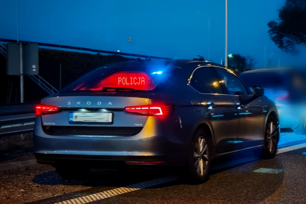 Radiowóz policyjny z włączonymi sygnalizatorami świetlnymi zatrzymany na drodze, wyświetlający napis "POLICJA".