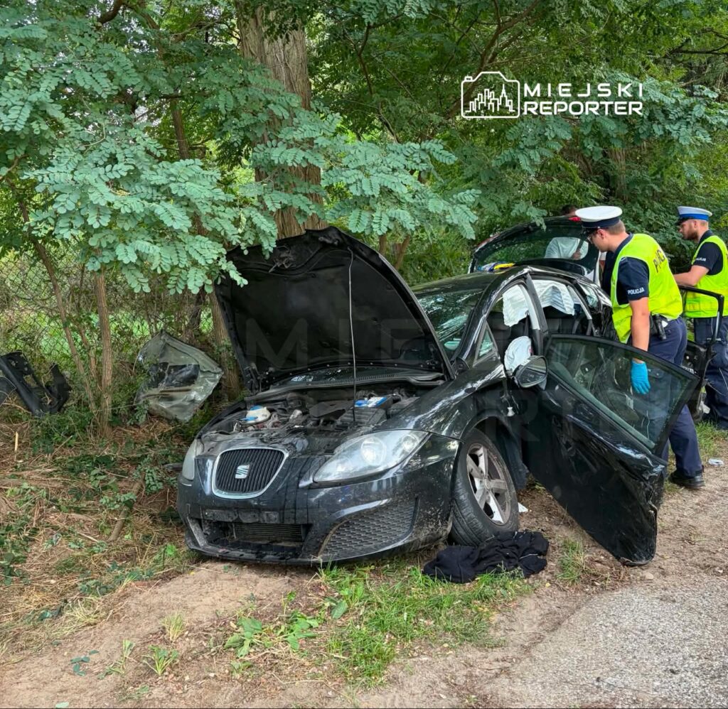 Czarny samochód osobowy z otwartą maską stoi przy drodze, obok dwóch policjantów badających sytuację.